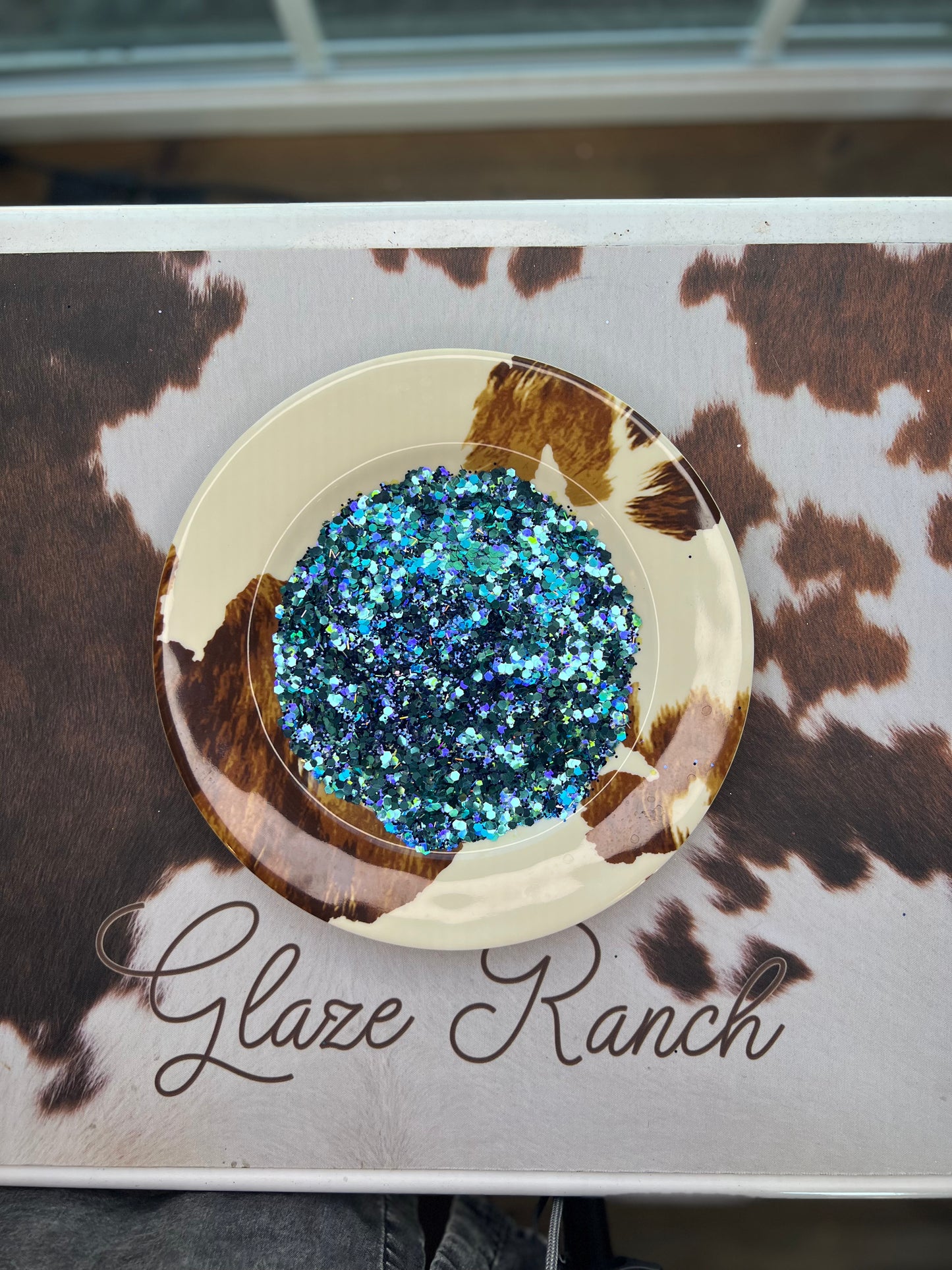 Decorative plate with blue glitter on a cow print background with 'Glaze Ranch' branding.