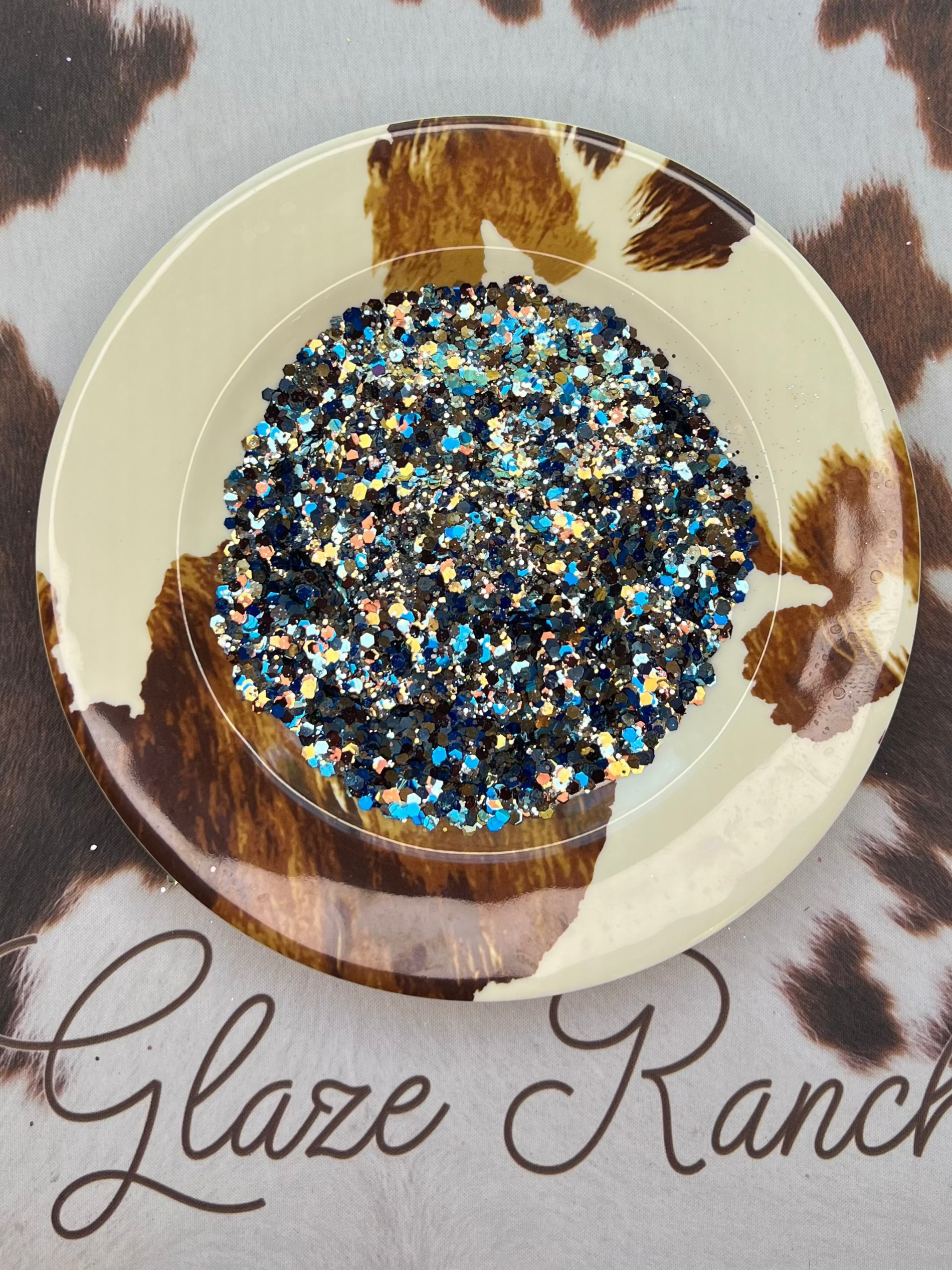 Decorative plate with blue and silver glitter on a cowhide-patterned surface with 'Glaze Ranch' branding.